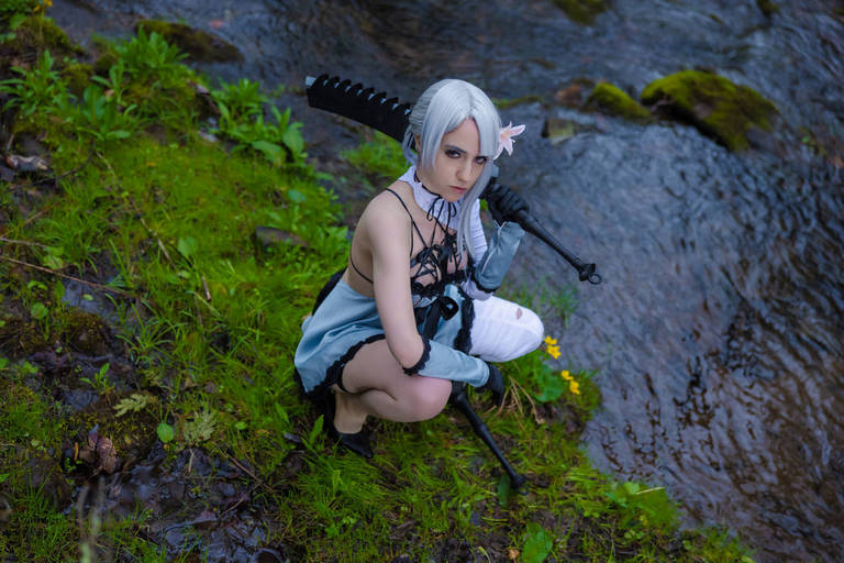 A person in cosplay with silver hair and a sword crouches by a stream surrounded by green grass and flowers.