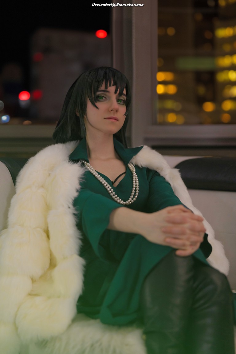 A person with dark hair and green eye makeup sits on a couch, wearing a green dress and a white faux fur coat, with pearl jewelry, in a dimly lit room with city lights in the background.