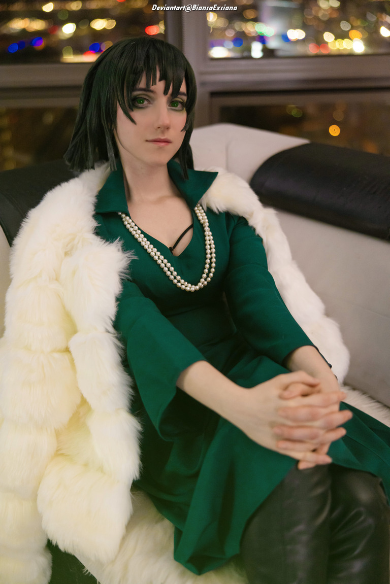 A person in a stylish green dress and a white fur coat, seated with hands clasped, showcasing a chic look against a cityscape background at night.
