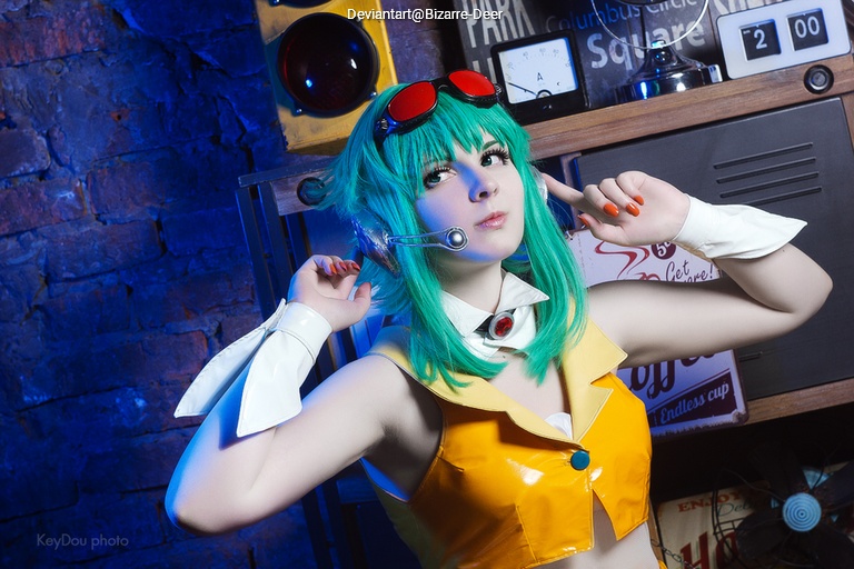 A person with green hair, wearing a yellow outfit with a white collar and red goggles, poses playfully while surrounded by retro decor.