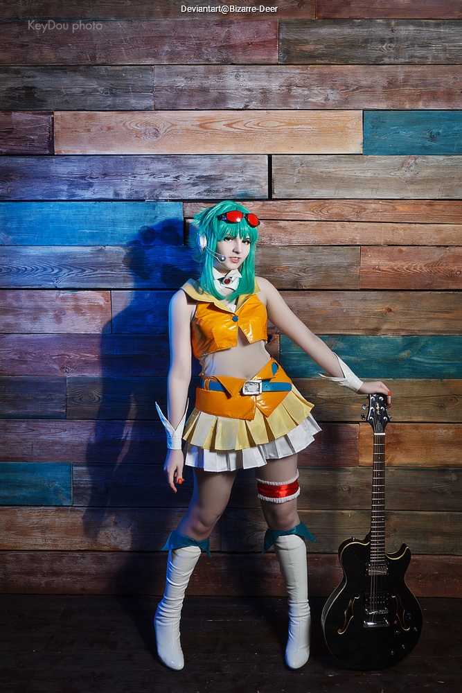 A person dressed in a colorful costume with a short skirt, wearing headphones and holding a guitar, standing in front of a wooden wall.