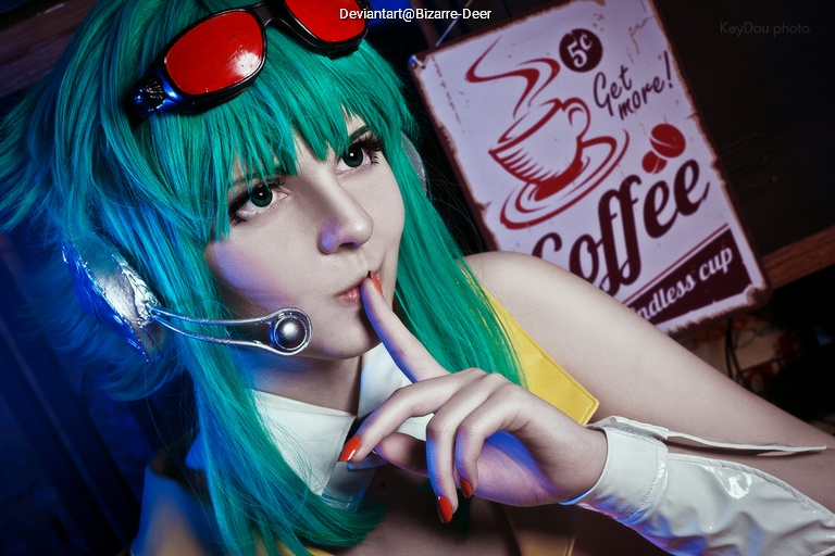 Person with green hair and red goggles, wearing a headset, holding a finger to their lips in a 'shh' gesture, against a coffee-themed backdrop.