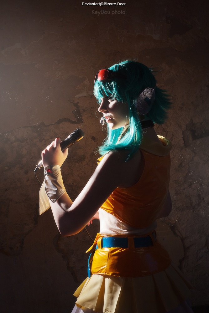 A person with teal hair wearing a yellow and orange outfit, holding a microphone, in a dimly lit environment with a textured wall.