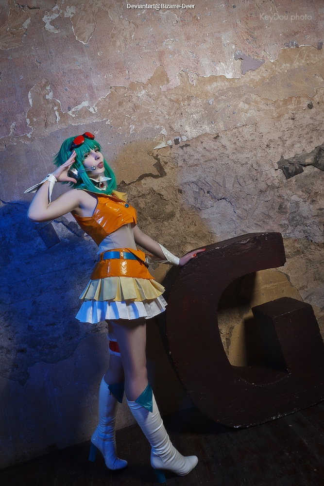 A person in a colorful costume with green hair and red accessories poses beside a large letter 'G' against a textured wall.