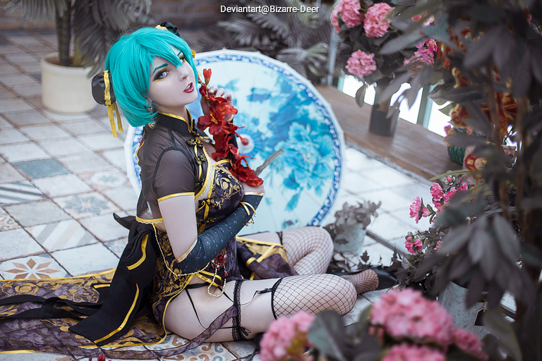 A person in cosplay with teal hair and intricate outfit sits among flowers, holding a red floral adornment and a decorative umbrella.