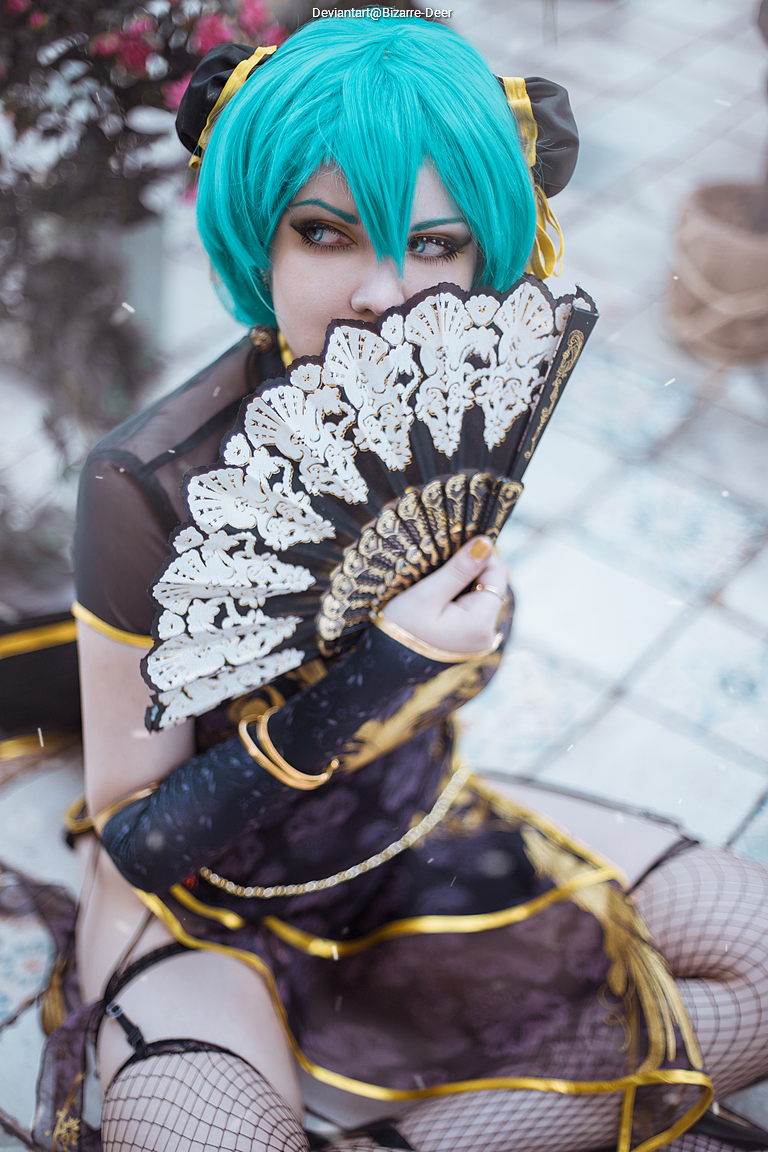 A person with turquoise hair wearing a black and gold outfit, holding a decorative fan. The background features floral elements and patterned flooring.