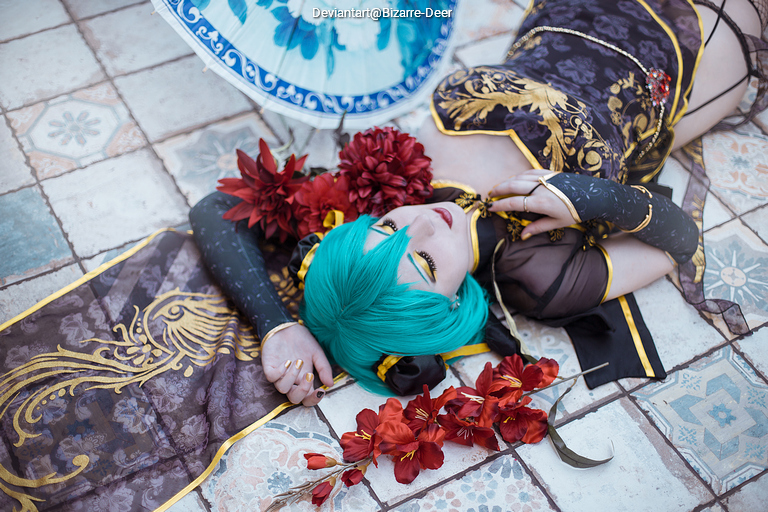 A person with turquoise hair dressed in a dark, embellished outfit lies on a patterned floor surrounded by red flowers and traditional decorative items.
