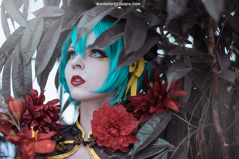A person with turquoise hair and dramatic makeup, adorned with large red flowers, gazes upward amidst lush green foliage.