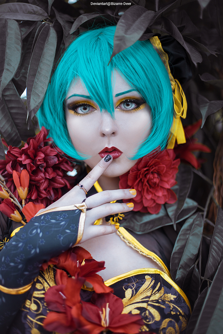 A person with turquoise hair and dramatic makeup surrounded by dark leaves and red flowers, posing with a finger to their lips.