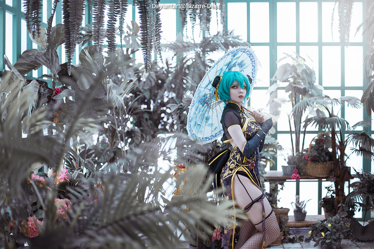 A person dressed in elaborate cosplay with turquoise hair and a decorative parasol, standing among lush plants and flowers in a greenhouse setting.