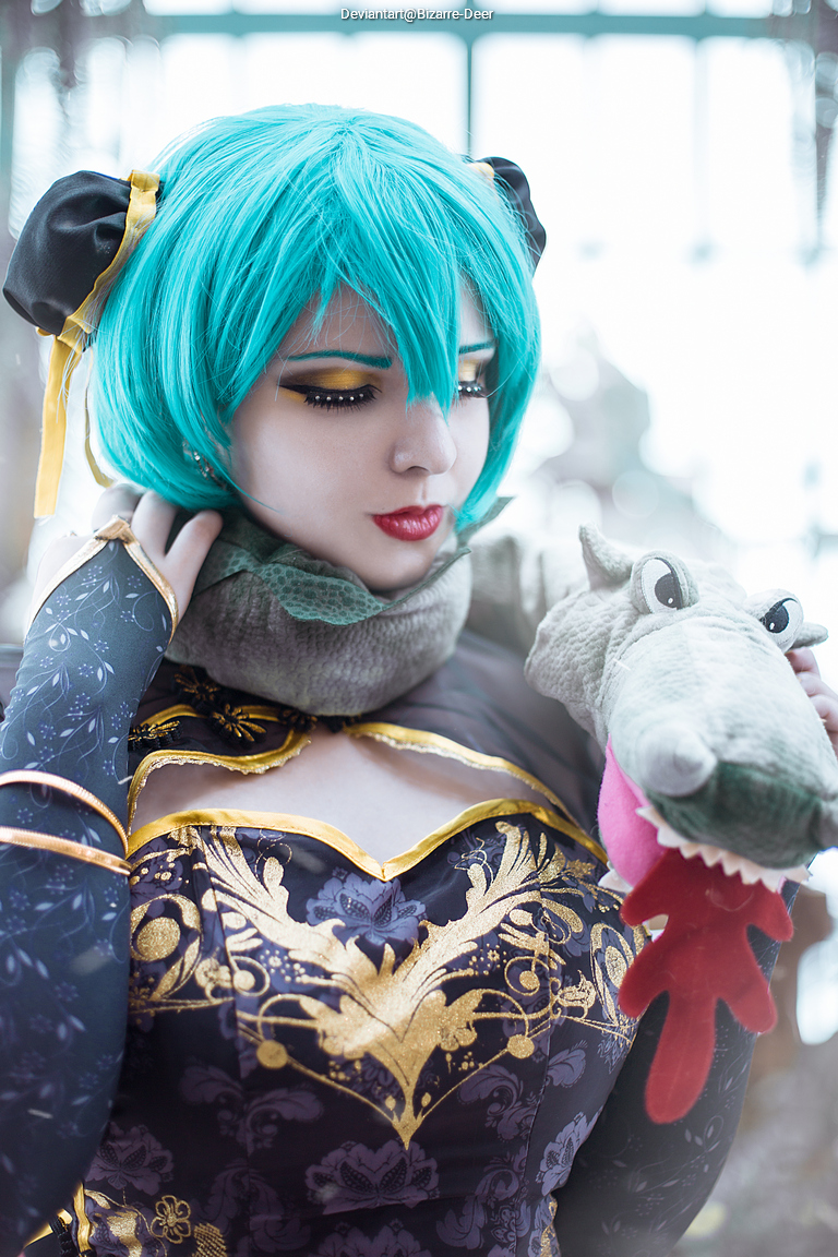 A person with turquoise hair wearing a decorative outfit featuring gold patterns and a scarf, holding a plush toy resembling a dragon.