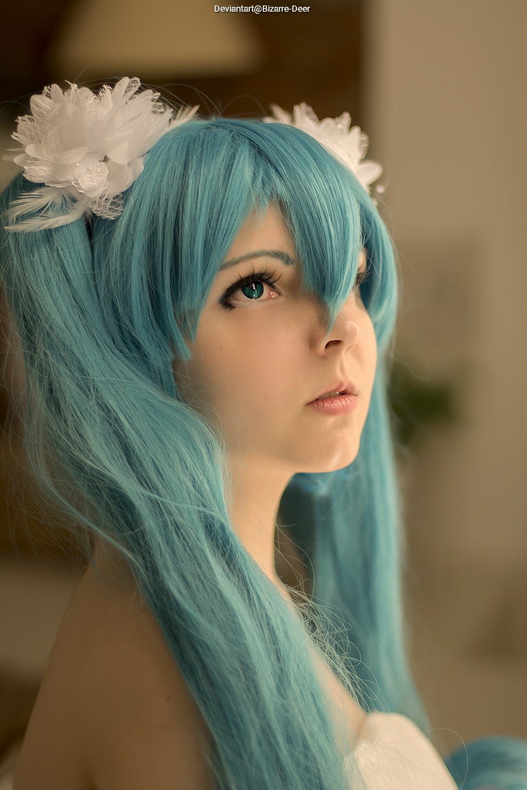 A close-up portrait of a person with long, blue hair styled in pigtails, wearing white flower accessories. The person has a neutral expression and is looking upwards, with a soft, blurred background.