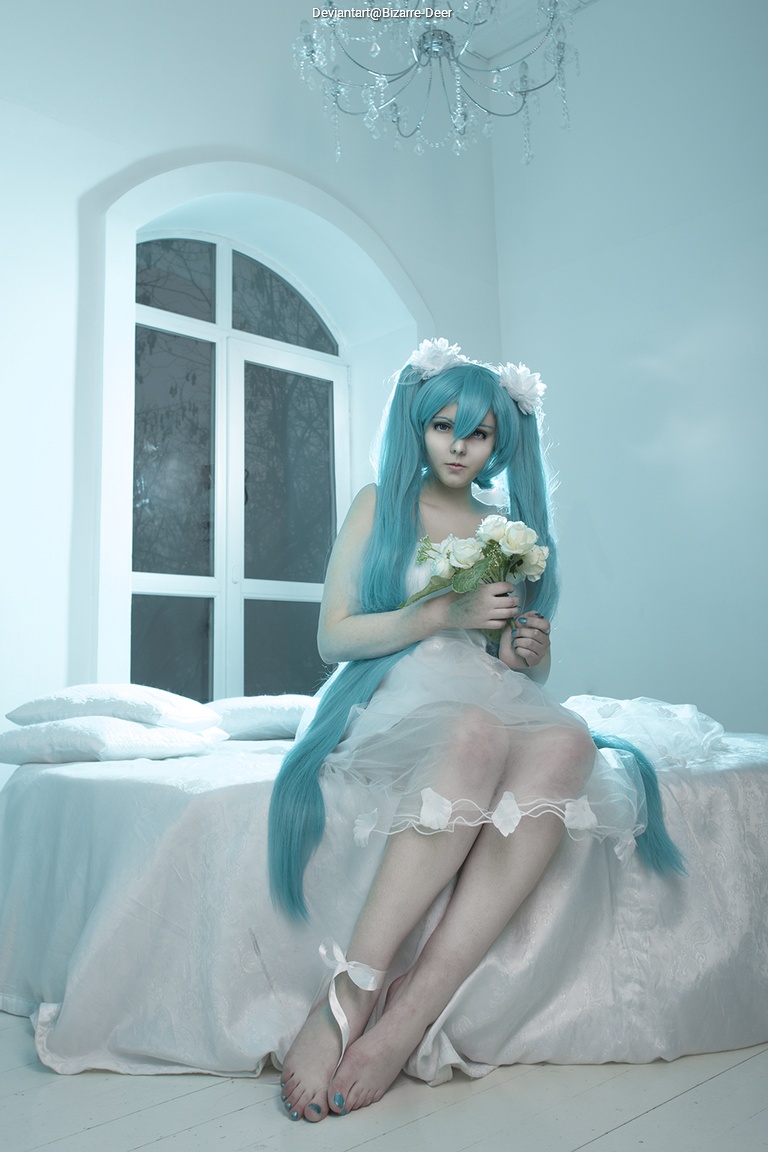 A person with long turquoise hair, wearing a white dress, sits on a bed holding a bouquet of flowers. The room is softly lit with white decor and a chandelier above.