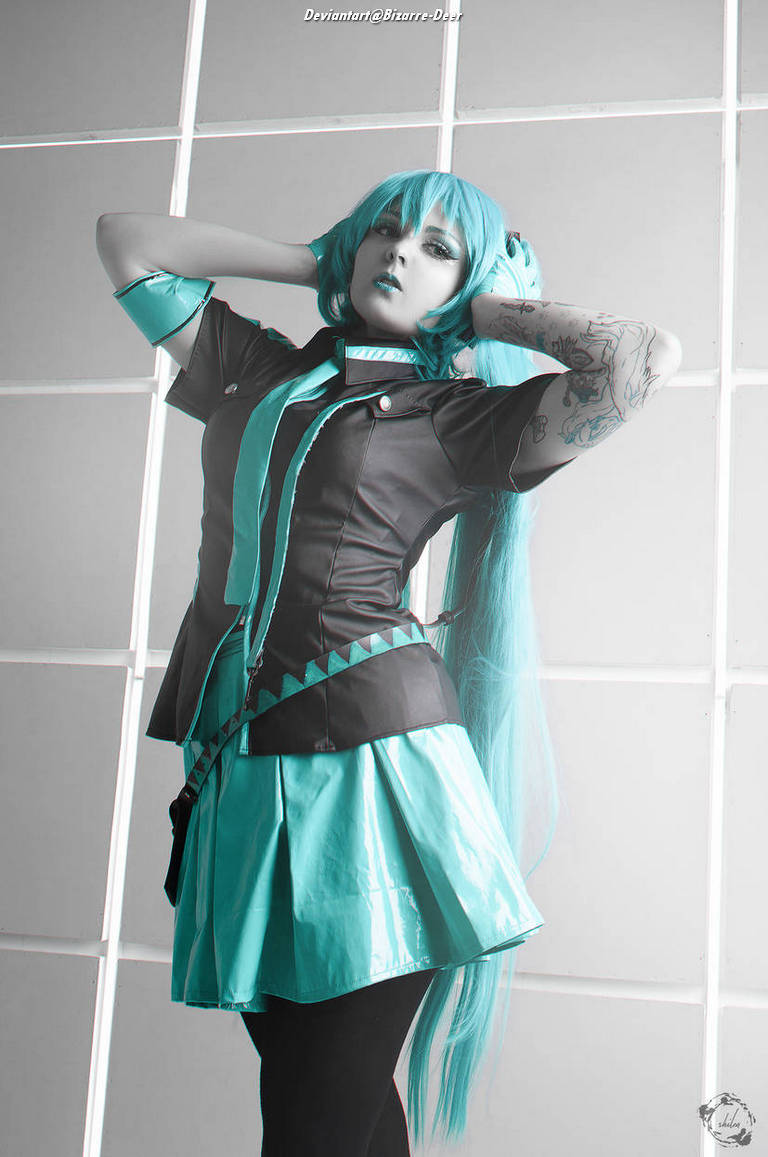 A person in a teal and black cosplay outfit striking a pose against a white backdrop, with long teal wig and tattoos visible on their arms.
