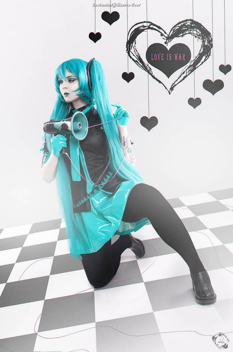 A person with long turquoise hair wearing a black and turquoise outfit, kneeling on a checkered floor, holding a megaphone. A heart-shaped graphic reading 'LOVE IS WAR' is in the background.