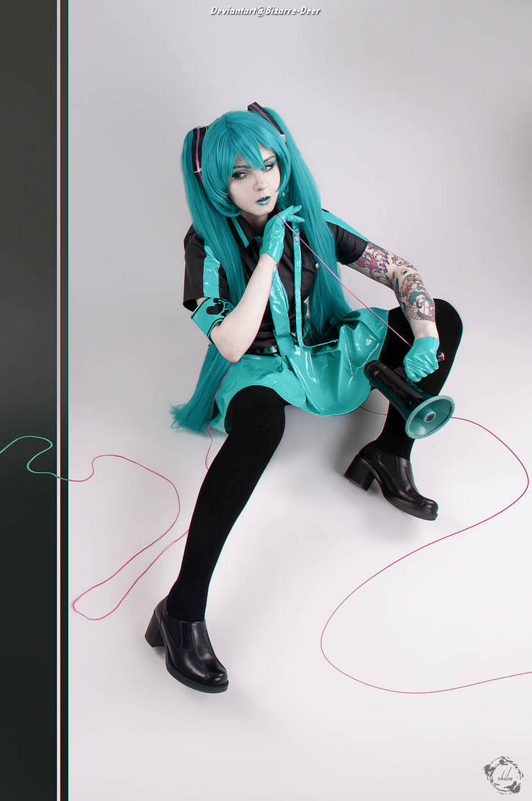 A person with long teal hair and dramatic makeup, dressed in a teal and black outfit, sitting on the floor while holding a spool of thread and posing with an expressive look. The background is minimalistic with a focus on the character's vibrant attire and styling.