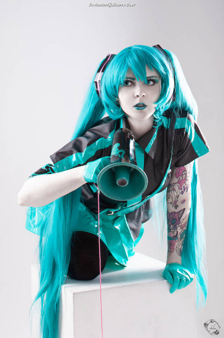 A person in a vibrant turquoise wig and outfit poses with a megaphone, showcasing a dynamic expression. The background is neutral, emphasizing the character's colorful costume and tattoos.
