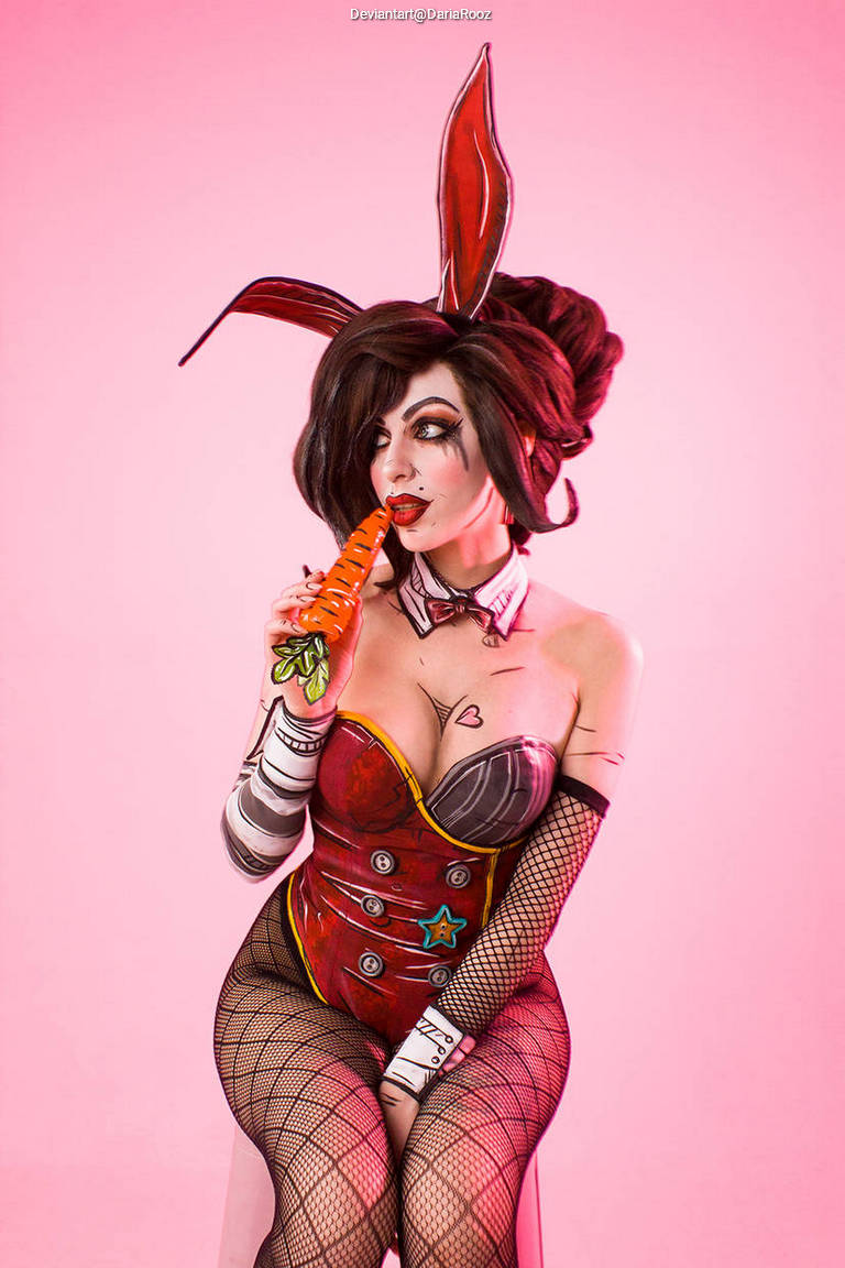 A person with a vintage bunny costume and dramatic makeup holds a carrot while sitting against a pink background.