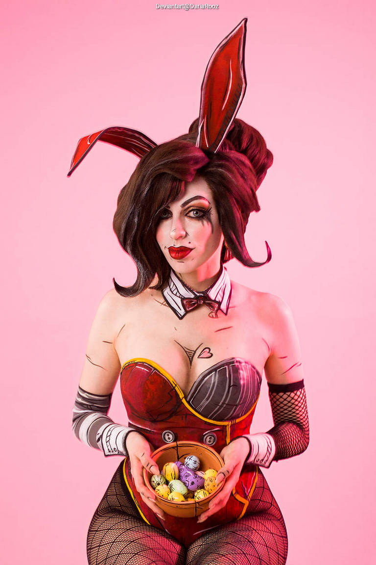 A woman wearing a bunny costume holds a bowl of colorful eggs, posing against a pink background.