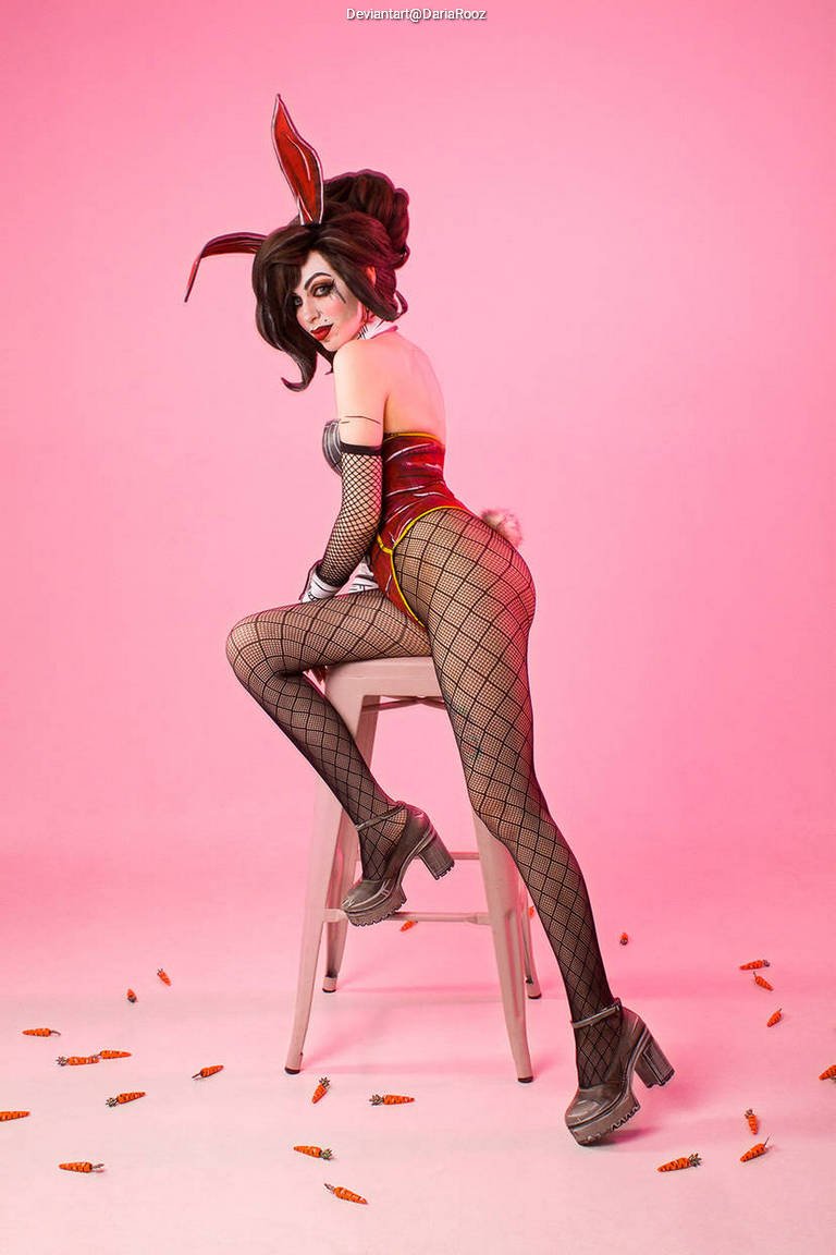 A person in a playful costume with bunny ears sits on a stool against a pink background, wearing fishnet stockings and high-heeled shoes, with small carrot decorations on the ground.