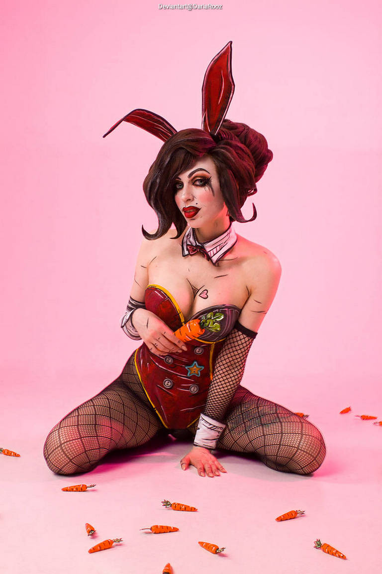 A character in a playful bunny costume with large ears, sitting on the floor surrounded by carrot props, against a pink background.