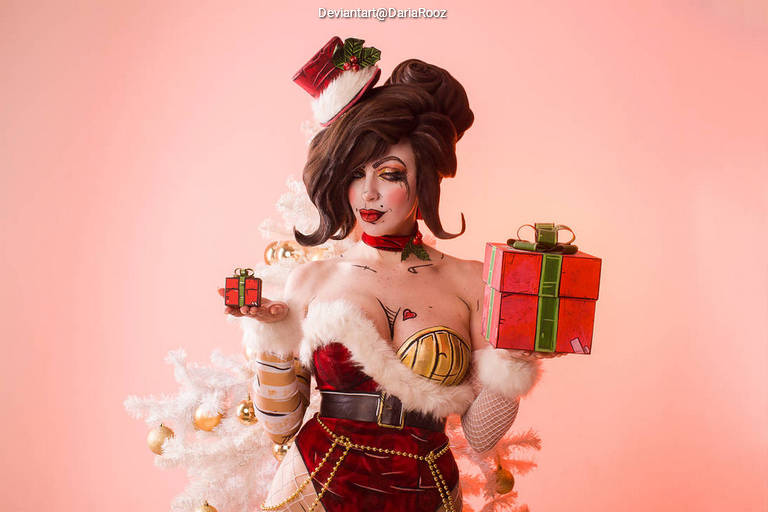 A person dressed in a festive holiday-themed outfit, featuring a red and white costume with accessories like a Santa hat, holding two gifts in front of a decorated Christmas tree, against a pink background.