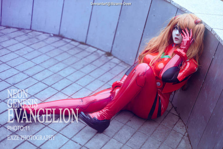 Cosplayer dressed as Asuka Langley Soryu from Neon Genesis Evangelion, sitting against a wall in a red outfit, portraying a thoughtful expression.