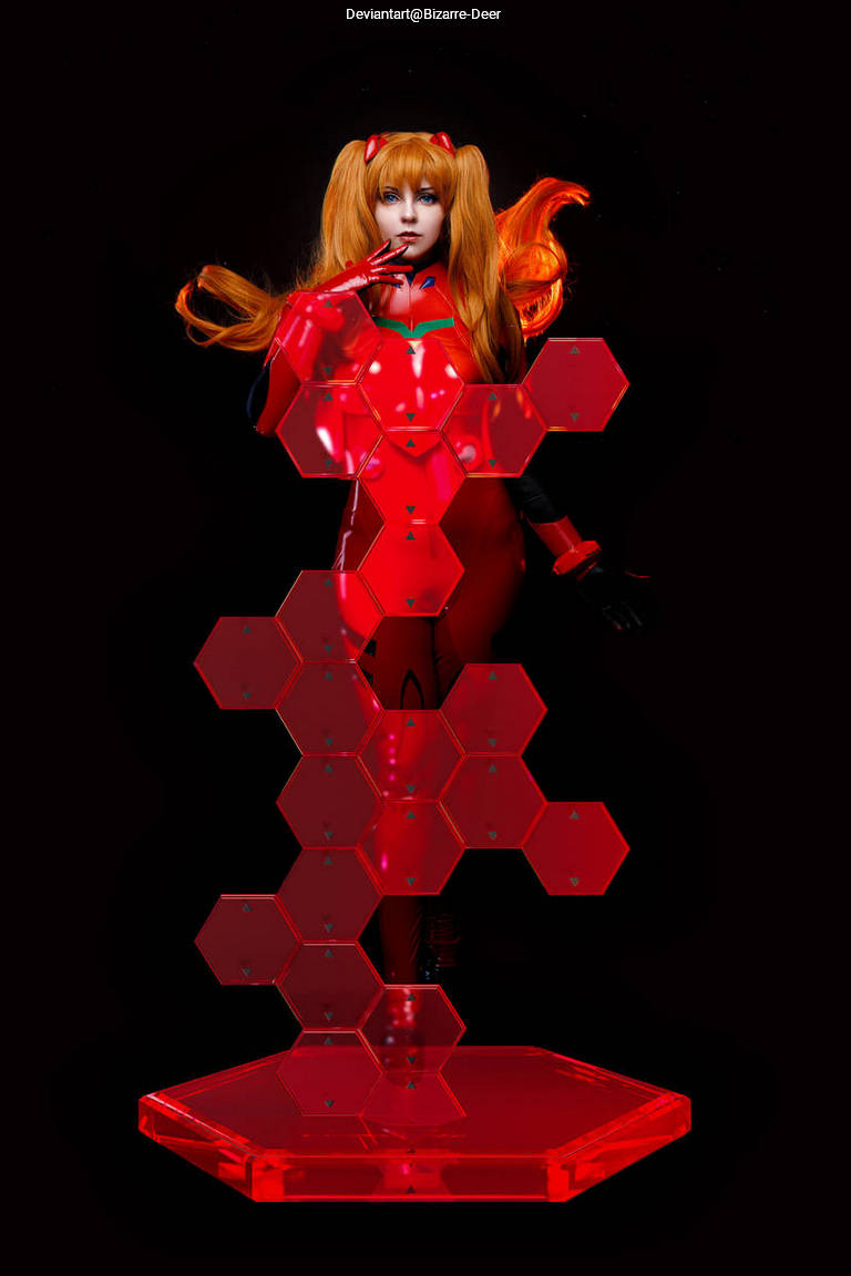 Cosplayer dressed as Asuka from the anime series Neon Genesis Evangelion, posed in a bright red suit with hexagonal display stand, against a black background.