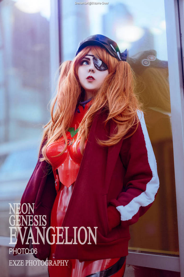 A person dressed as Asuka Langley Soryu from Neon Genesis Evangelion, showcasing vibrant orange hair and a stylish outfit, standing in front of a glass window with a cityscape background.
