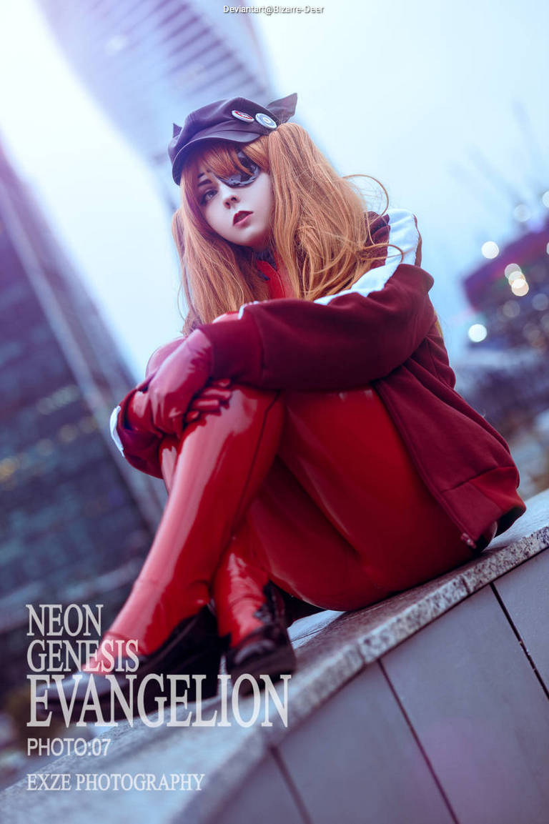 A person dressed in a detailed cosplay of Asuka Langley Soryu from Neon Genesis Evangelion, sitting on an outdoor ledge in urban surroundings, with a soft blue and gray background.