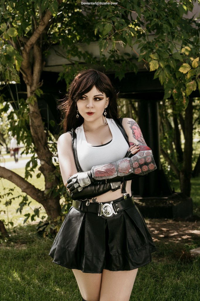 A person in a cropped white tank top and black leather skirt stands outdoors, crossing their arms. They have long brown hair, striking makeup, and are wearing decorative gloves with red accents. The background features leaves and a structure.