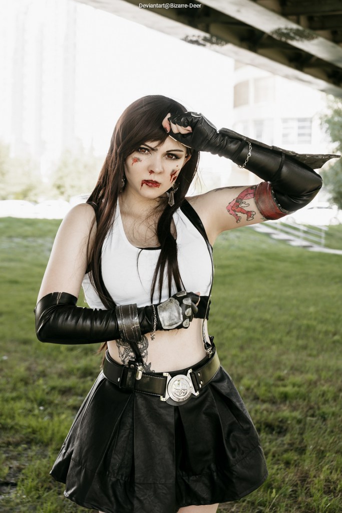 A person in a costume resembling a character, featuring a white top, a black skirt, and black gloves, with facial makeup suggesting a battle theme and a serious expression, standing outdoors under a structure.
