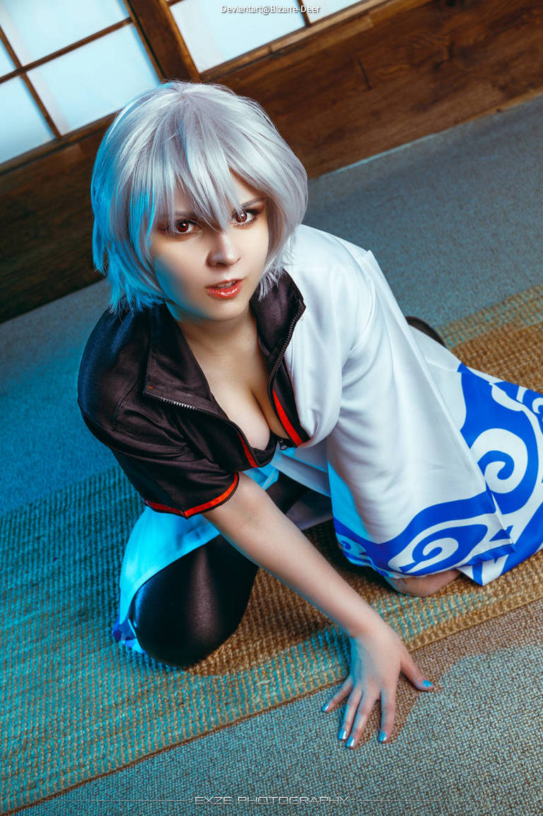 A person with short, silver hair in a stylized outfit, crouching on a mat in a traditional indoor setting, with wooden panels and soft lighting.
