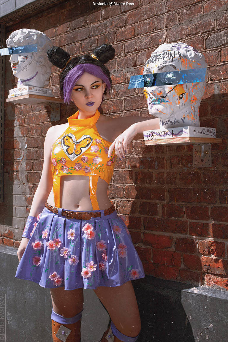 A person with purple hair styled in buns poses against a brick wall, wearing a colorful outfit with floral patterns and a butterfly design. Two art sculptures of heads are displayed on the wall, partially obscured by sunglasses and graffiti.
