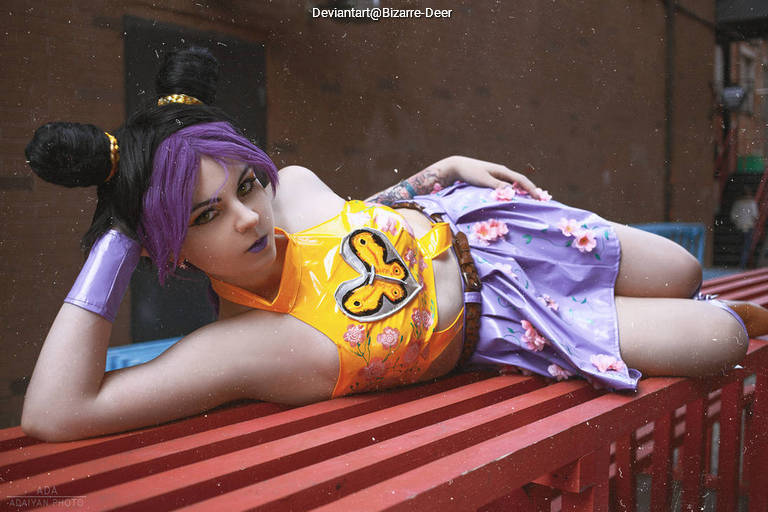 A person with purple hair poses on a red surface, wearing a shiny yellow top with a butterfly design and a floral purple skirt. They have distinctive purple and black bun hairstyles and are showcasing a tattoo on their arm.