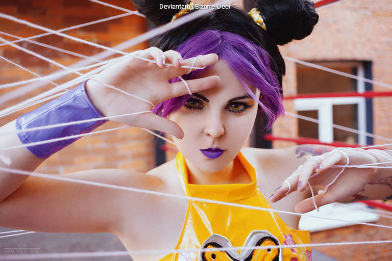 A person with purple hair and striking makeup poses dramatically, surrounded by white string, suggesting a theme of entrapment or performance art.