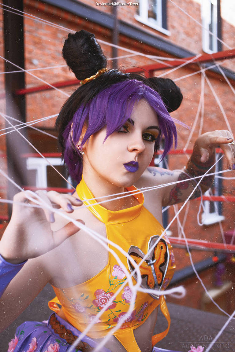 A person with purple hair styled in buns is posing with a serious expression. They are wearing a bright yellow outfit with floral designs and a butterfly motif. The background features red structures and a web-like material around them.