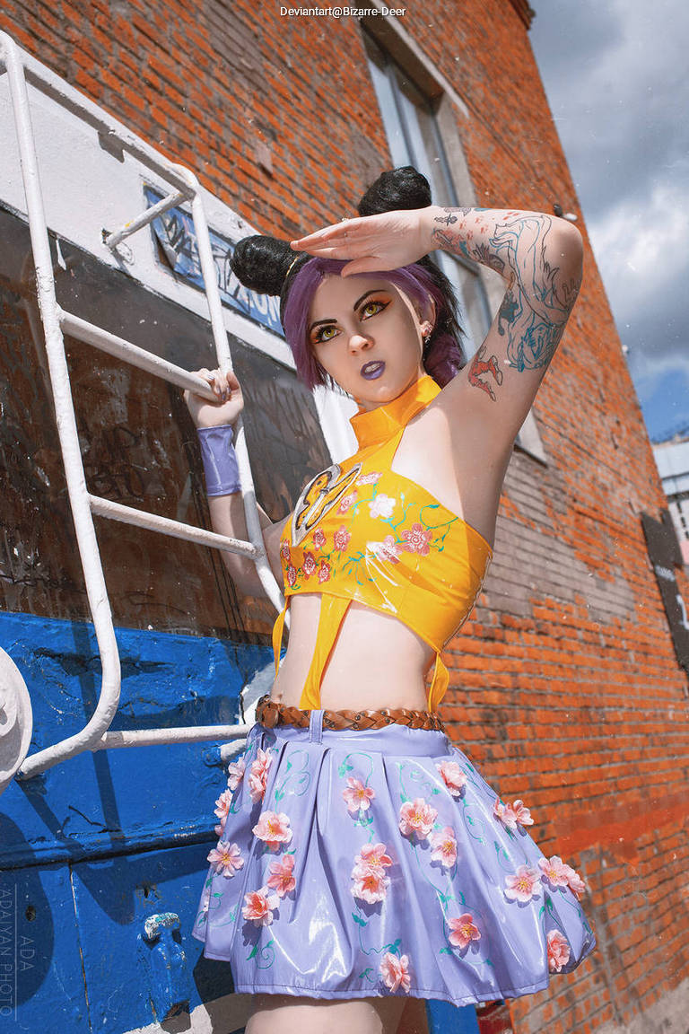 A person with purple hair and mouse ear accessories poses confidently in front of a brick wall, wearing a colorful outfit with floral designs.