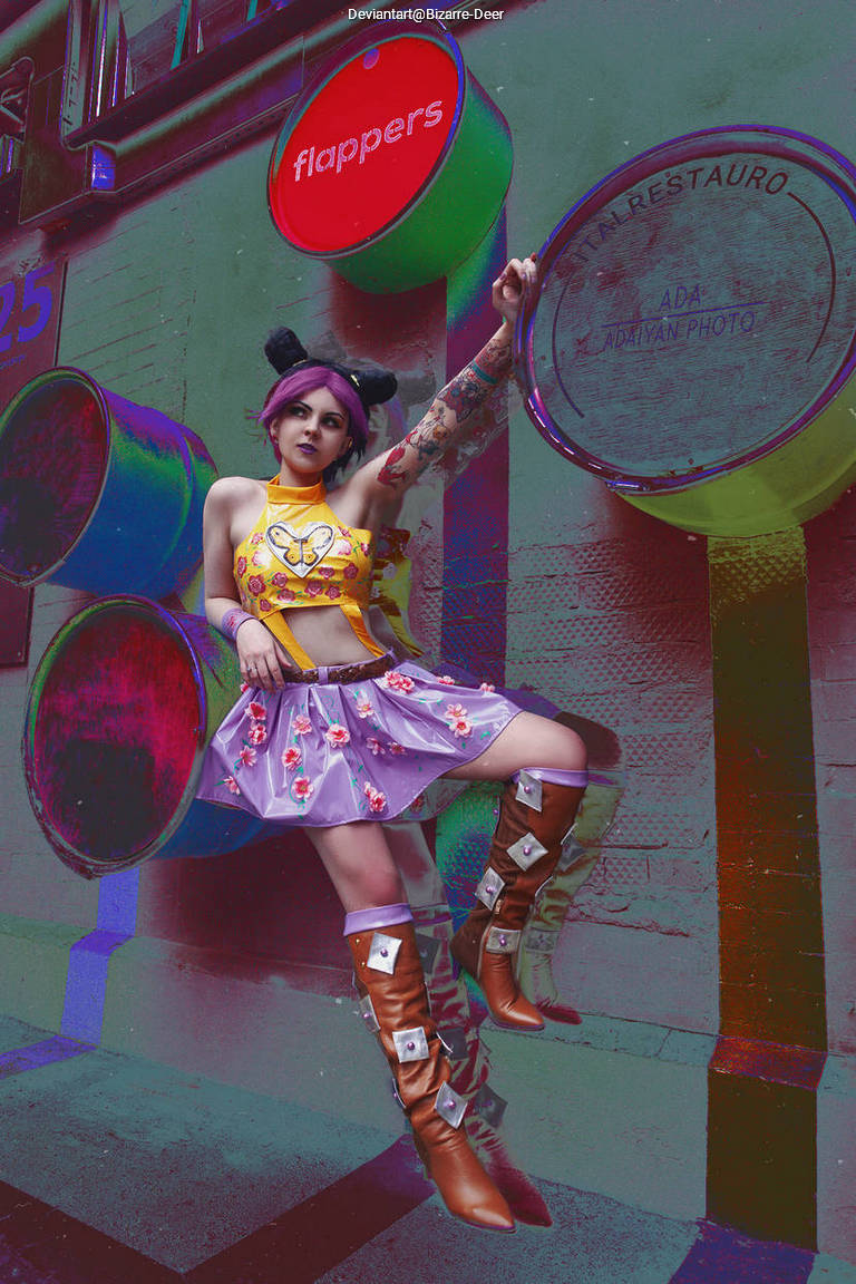 A person with purple hair and bunny ears poses in a colorful outfit featuring a butterfly design and a purple skirt, against a vibrant urban backdrop with barrels and signage.