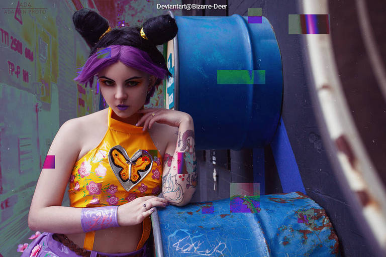 A person with purple hair styled in buns poses beside a blue barrel, wearing a colorful outfit with floral patterns and a butterfly design.