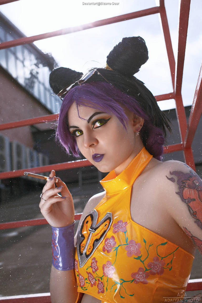 A person with purple hair styled in a bob, wearing black cat ears, bold yellow make-up, and an orange outfit adorned with butterfly and floral designs. They are holding a cigarette and posing confidently.