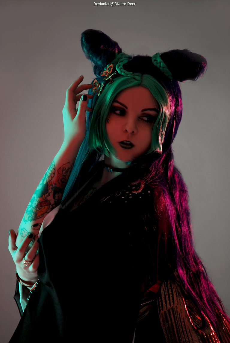 A person with green and black hair styled in a unique way, wearing a dark outfit with decorative elements, posing with an expressive look against a softly lit background.