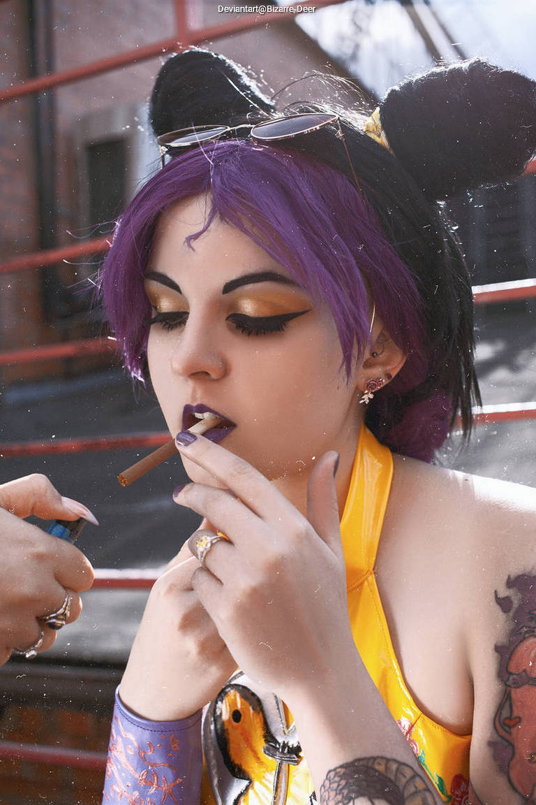 A person with purple hair styled in pigtails, wearing a vibrant yellow outfit with tattoos visible on their arm, is holding a cigarette with a focused expression. They have dramatic eye makeup and are surrounded by urban scenery.