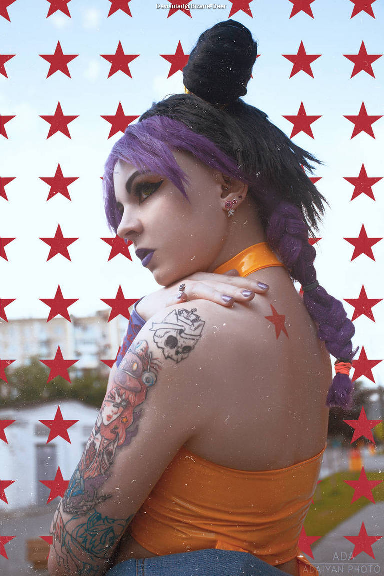 A woman with purple hair styled in a braid and bun, wearing an orange top, poses against a background of red stars. She has multiple tattoos visible on her shoulder and arm.