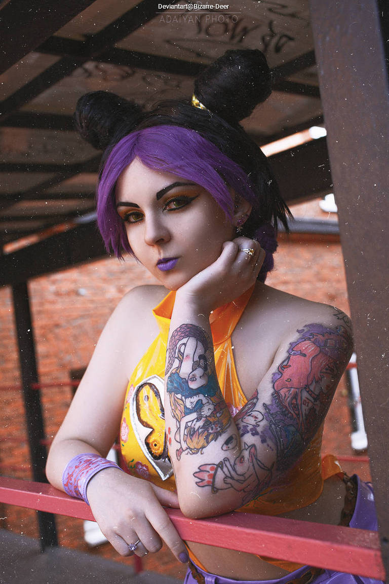 A person with purple hair styled in two buns, wearing a bright orange top, poses with tattoos visible on their arms against a rustic backdrop.