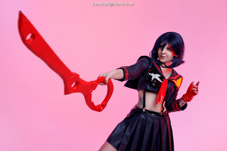 A person in a black outfit with red accents, holding a large red weapon, posing against a pink background.