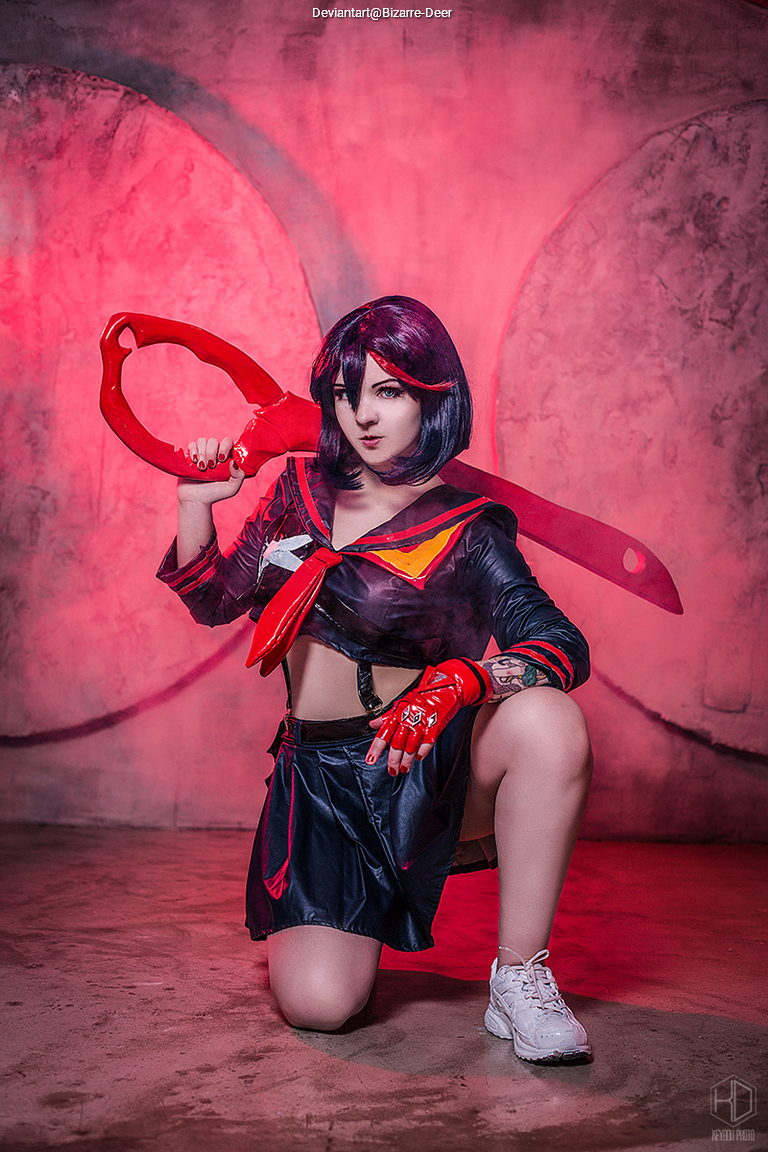 A cosplayer dressed as a character with dark hair and a red and black outfit, holding a large red weapon, posing in a kneeling position against a pink and gray backdrop.