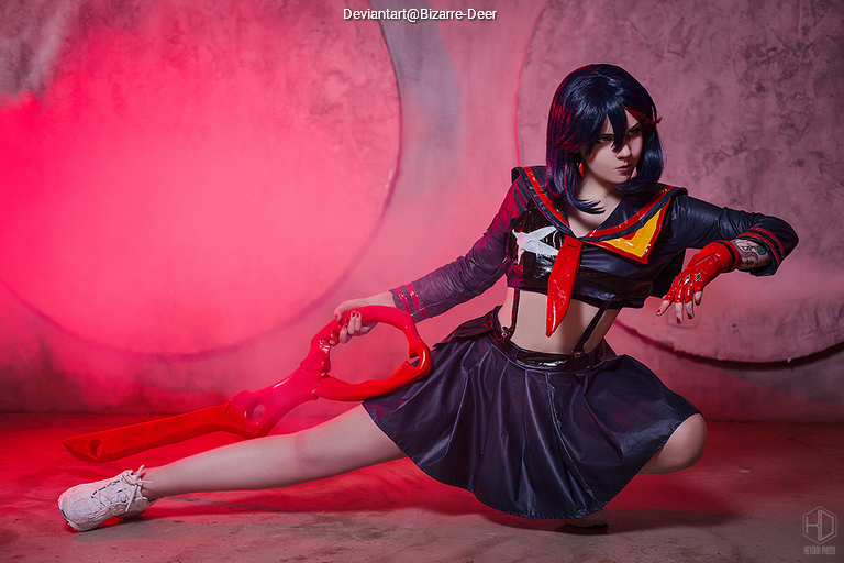 A female cosplayer in a black and red outfit, inspired by a character from an anime, striking a dynamic pose with a large red weapon in a dramatic setting with pink lighting.