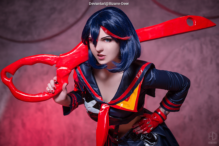 Cosplayer posing with a large red scissor blade, wearing a black and red outfit with a sailor-style top, featuring blue hair and dramatic makeup.