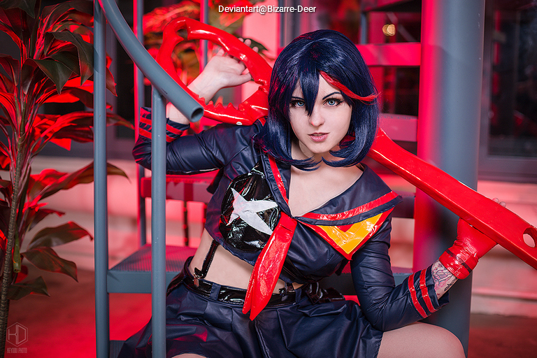 A cosplayer dressed as a character with blue hair, wearing a black and red outfit, poses dramatically on a staircase with red lighting.